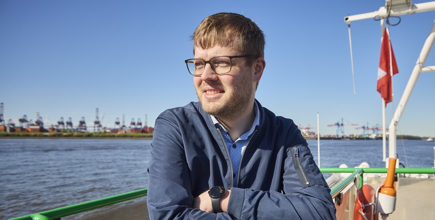 Man with glasses leaning on a ferry’s railing, looking out over a port with water and container cranes in the background.