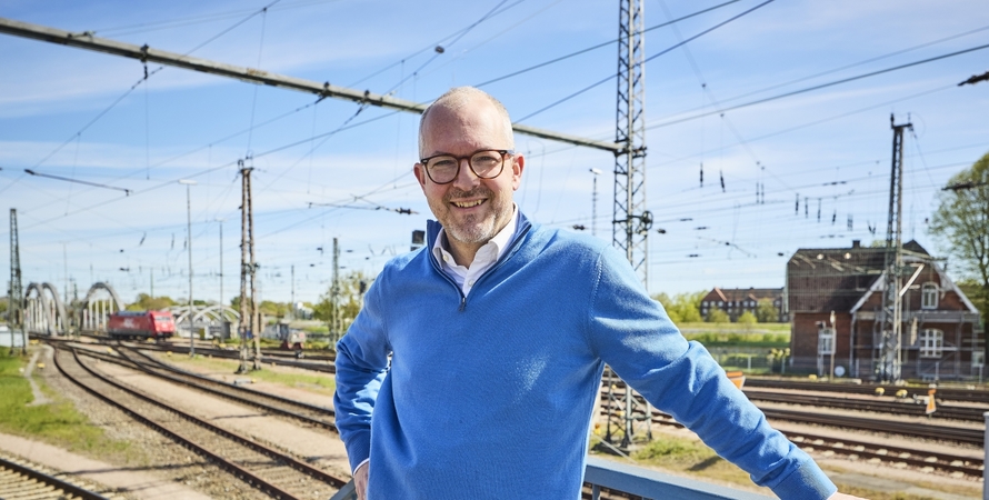 Man in a blue sweater standing on a platform in front of railway tracks at a terminal, looking at the camera.