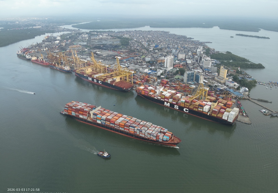 Aerial view of the container terminal in Buenaventura, Colombia, with multiple container vessels berthed along the quay, container stacks and ship-to-shore cranes handling cargo operations. The surrounding waterfront cityscape and waterways are visible in the background.