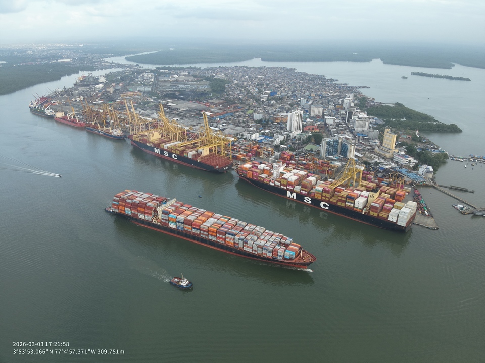 Aerial view of the container terminal in Buenaventura, Colombia, with multiple container vessels berthed along the quay, container stacks and ship-to-shore cranes handling cargo operations. The surrounding waterfront cityscape and waterways are visible in the background.